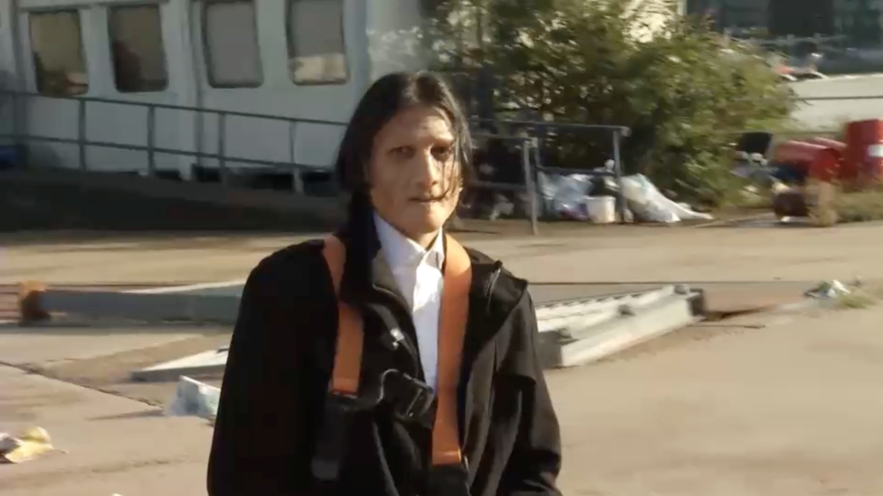 Um homem com longos cabelos negros usando uma prótese no rosto e vestido com um casaco escuro com uma camisa branca abotoada no colarinho.