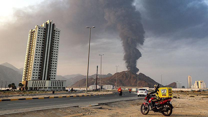 سائق دراجة نارية متوقف على جانب الطريق السريع وفي الخلف الدخان يتصاعد من المدينة الصناعية في الفجيرة خلف تلة من تراب
على يمين الطريق مبنى سكني باللون الأبيض والبني