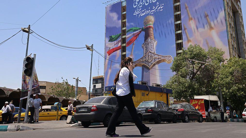 امرأة تمر أمام لافتة عملاقة تصور صواريخ إيرانية وسيفًا للإمام علي، الإمام الأول للشيعة، في ساحة فاناك في طهران في 10 يونيو/حزيران 2026. 