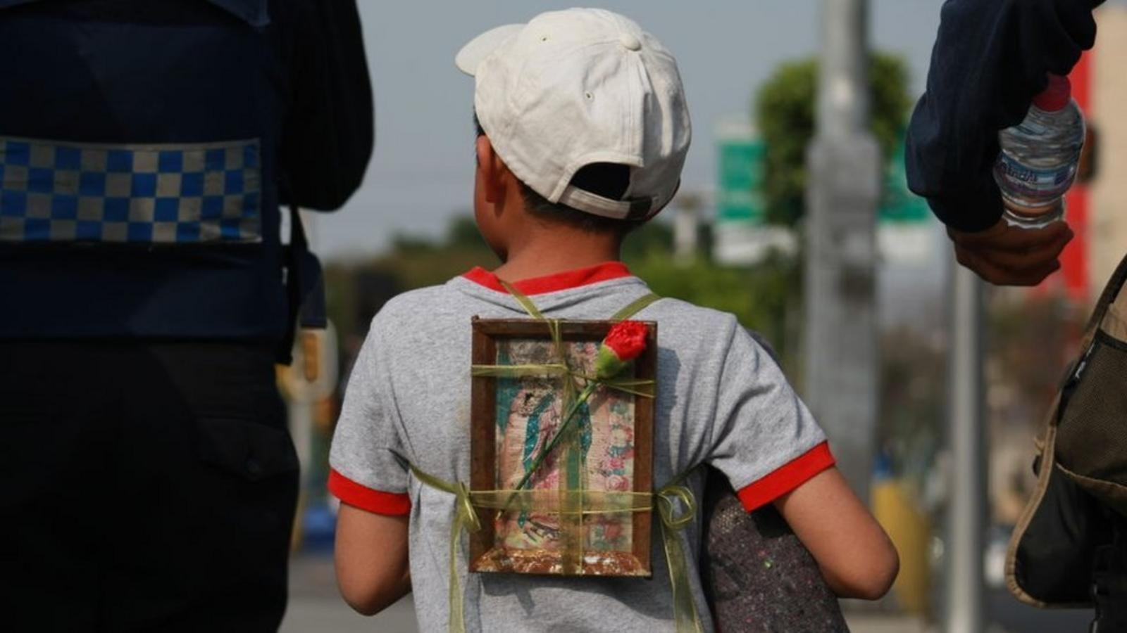 Un niño con una gorra blanca y playera gris con bordes rojos, de espaldas a la cámara y flanqueado por dos adultos, lleva la imagen de la Virgen de Guadalupe atada a su espalda