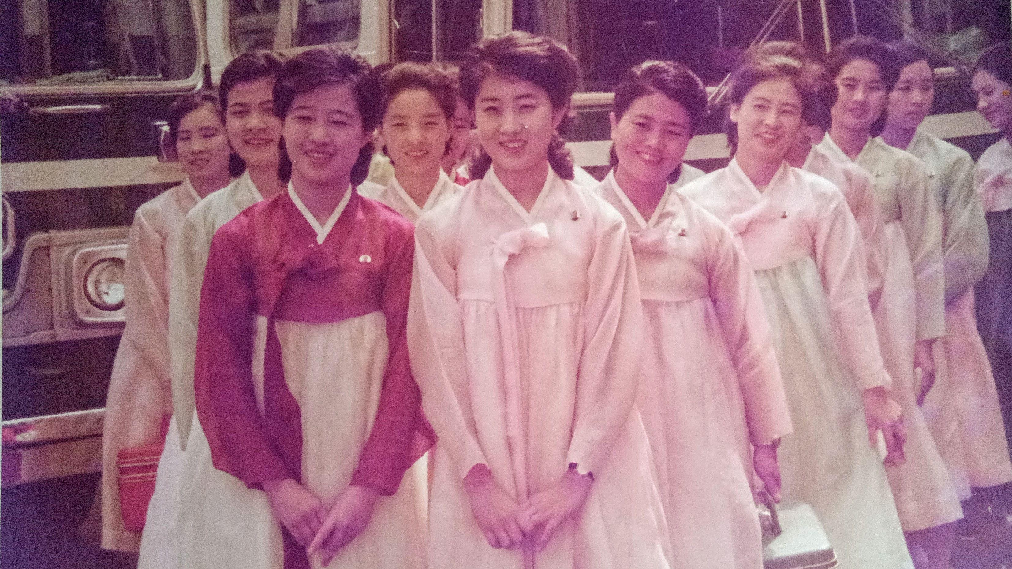 Cerca de uma d&uacute;zia de jovens com vestidos tradicionais coreanos est&atilde;o na frente dos ve&iacute;culos. Ko Yong-hui est&aacute; na frente, &agrave; esquerda 