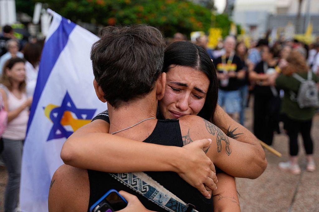 Una mujer se ve de frente abrazando a lo que parece ser un hombre, de espaldas. Ella tiene los ojos cerrados y un gesto que parece indicar a la vez alivio y tristeza. El hombre de espaldas va con una camiseta de tirante ancho negra y tiene varios tatuajes en su brazo derecho. Al fondo se ve una bandera de Israel que, en el centro, tiene entrelazada en la estrella de David un lazo amarillo, símbolo de la petición de liberación de los rehenes. Al fondo, difuminados, se ve mucha más gente en la plaza.