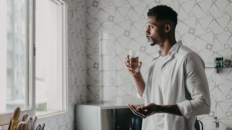 Homem se prepara para tomar um comprimido, segurando um copo d'&aacute;gua e olhando para a janela