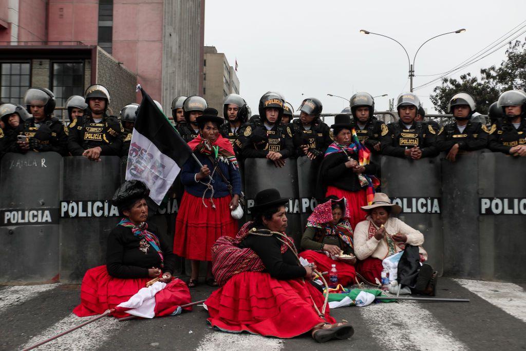 Manifestantes indígenas, con la vestimenta tradicional de cholita, de falda larga roja con enaguas blancas, chaqueta negra y sombrero tipo hongo negro, sentadas en el suelo rodeadas de efectivos policiales con escudos antidisturbios.