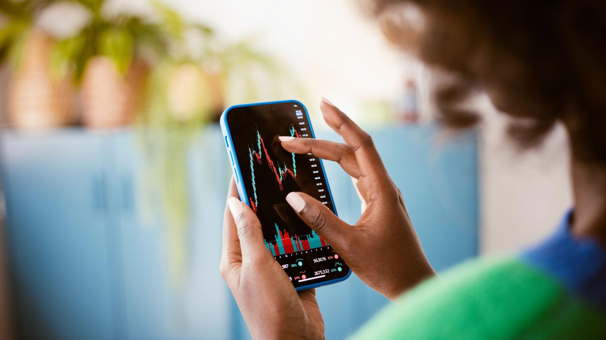 Mulher vista de costas com celular na mão, analisando gráficos de investimentos na tela do aparelho
