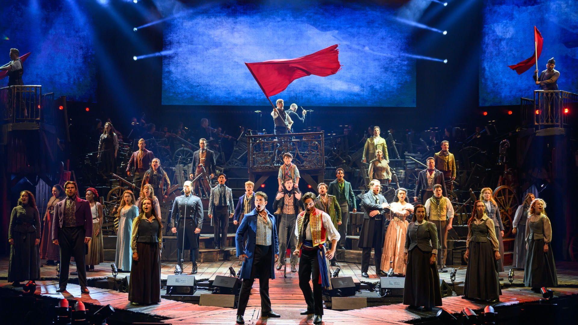 El elenco actuando durante la rueda de prensa de Los Miserables en Sidney, Australia. El elenco, vestido con trajes que recuerdan a la ropa de la época de la Revolución Francesa, se coloca en formación sobre el escenario. Al fondo, un hombre ondea una bandera roja y se encuentra de pie sobre una recreación de las barricadas que se utilizaban como defensa durante los combates.