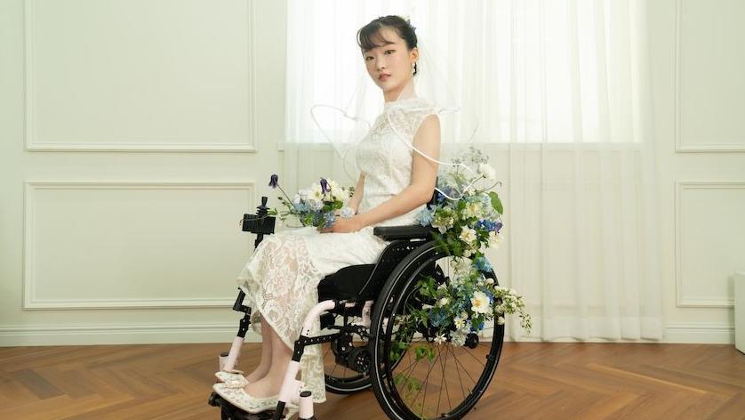 한 여성이 흰 웨딩드레스를 입고 휠체어에 앉아 있다. 휠체어에는 초록색, 흰색, 파란색 꽃들이 장식되어 있다.