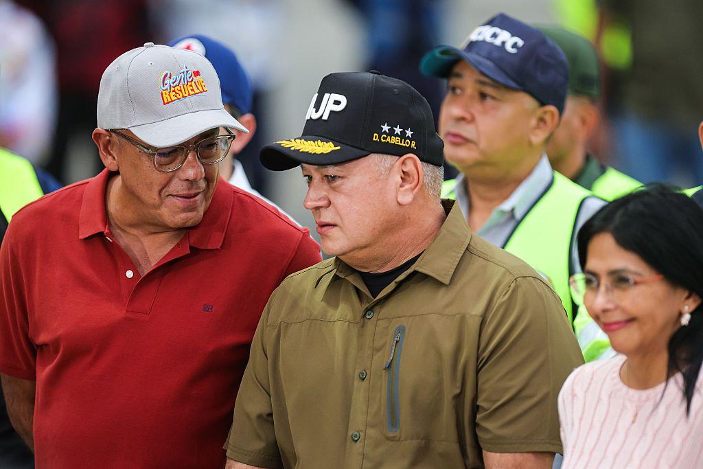 Jorge Rodríguez, a la izquierda, vestido con oolo rojo y gorrra gris, parece decirle algo a a Diosdado Cabello, en el centro, con camisa color militar y gorra negra y, en la esquina a la derecha, Delcy Rodríguez, que sonríe. 