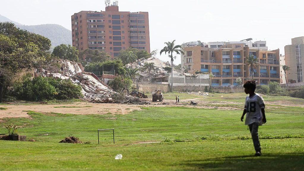 Edificios demolidos y dañados se observan detrás del Club de Golf y Yate Caraballeda mientras continúan las labores de rescate. Se ve un niño caminando sobre el césped. 