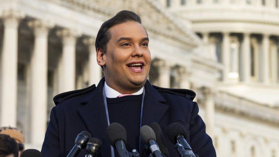 George Santos sorrindo ao falar diante de vários microfones em frente ao Capitólio americano
