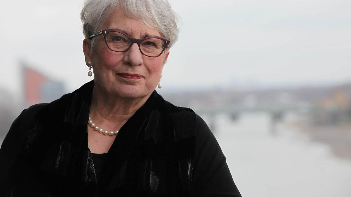 A professora Dame Uta Frith aparece em p&eacute; diante de um rio. Ela tem cabelos grisalhos curtos, usa &oacute;culos, colar de p&eacute;rolas e um casaco escuro