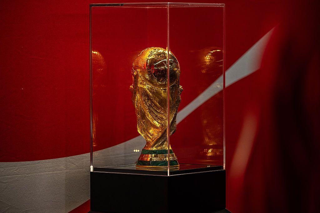 كأس العالم الذهبي بخطين بلون أخضر أسفل الكأس داخل زجاج للعرض والخلفية حمراء بخط أبيض.