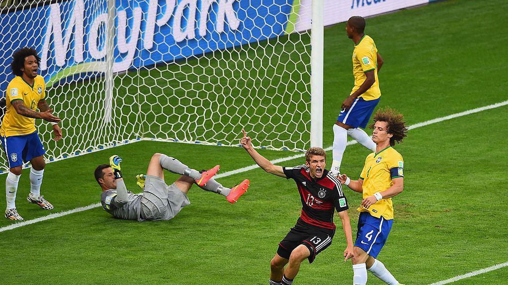 A imagem mostra um lance dentro da &aacute;rea durante uma partida entre Brasil e Alemanha. Enquanto um jogador alem&atilde;o comemora de bra&ccedil;os erguidos, jogadores brasileiros demonstram frustra&ccedil;&atilde;o ao redor da pequena &aacute;rea, com o goleiro ca&iacute;do no gramado segurando a bola e defensores observando a cena pr&oacute;ximos &agrave; linha do gol.