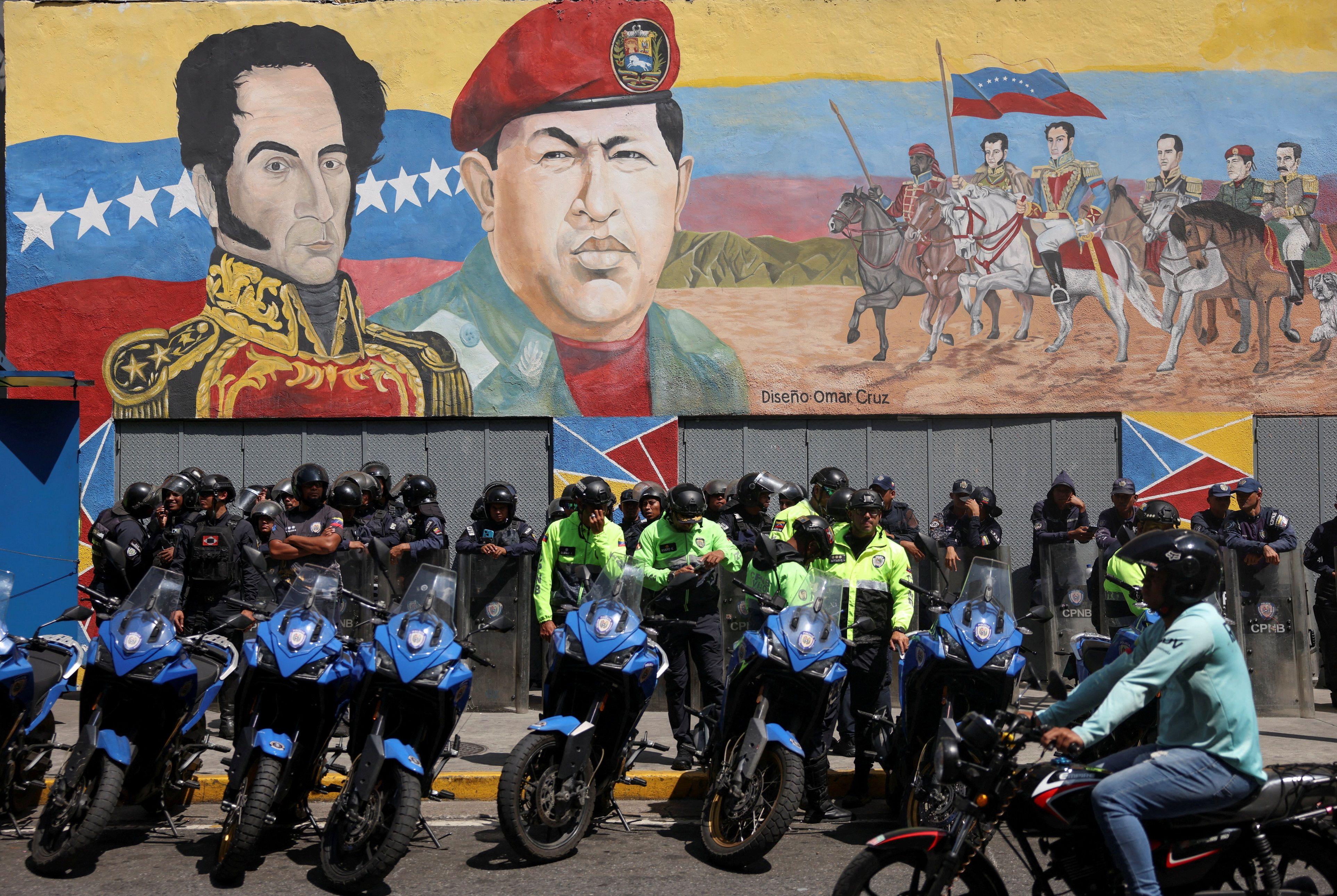 Fileiras de policiais de p&eacute; atr&aacute;s de motos, usando capacetes em frente a um enorme mural pintado de Sim&oacute;n Bol&iacute;var e Hugo Ch&aacute;vez