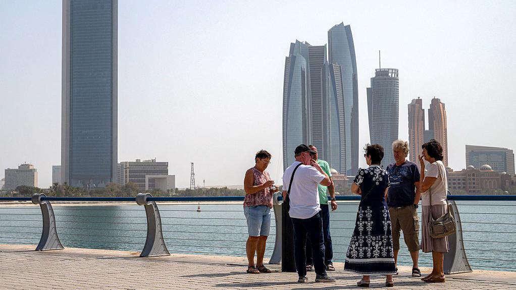 سياح رجال ونساء على رصيف شاطئ في أبو ظبي وفي الخلفية أبنية فخمة شاهقة