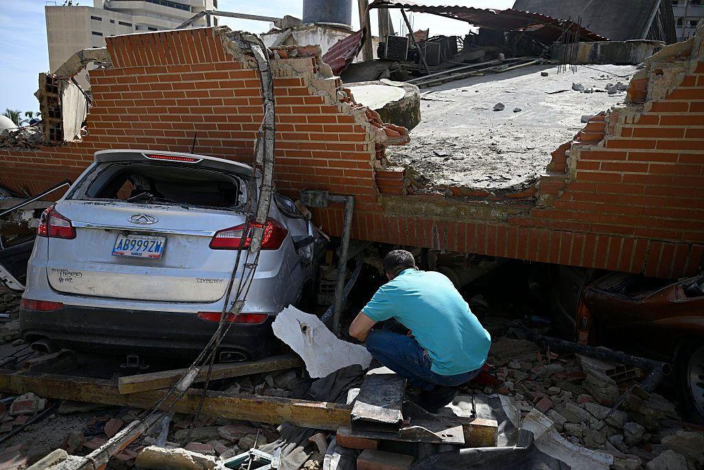 Un hombre acuclillado revisa el suelo alrededor de un auto aplastado por un pedazo de edificio que le cayó encima en los terremotos. 