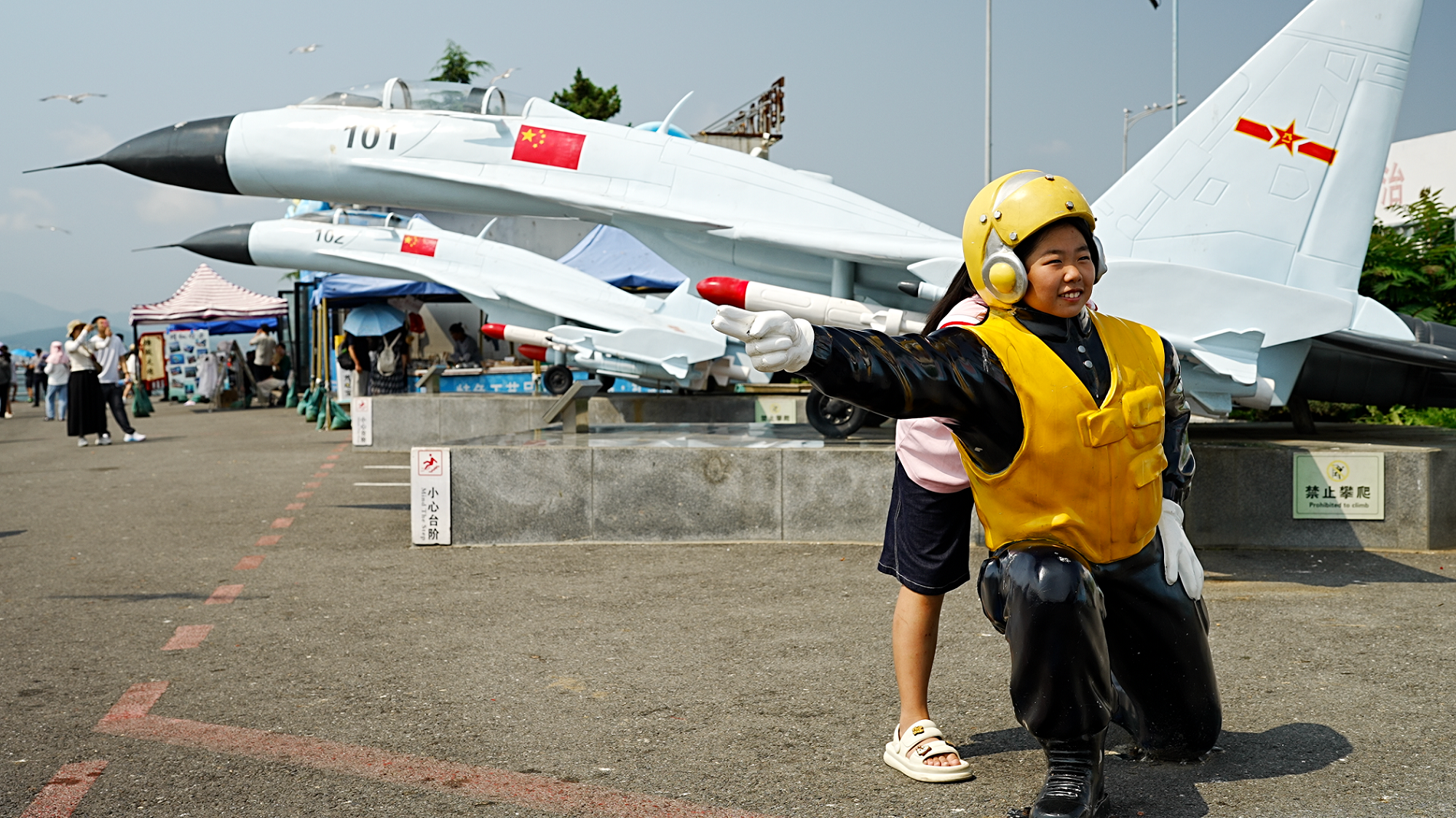 A girl in demin shorts and a pink shirt stand behind the statue of an airman. which is kneeling on the ground and pointing an arm in one direction- she puts her face above the statue's shoulders and smiles at the camera.