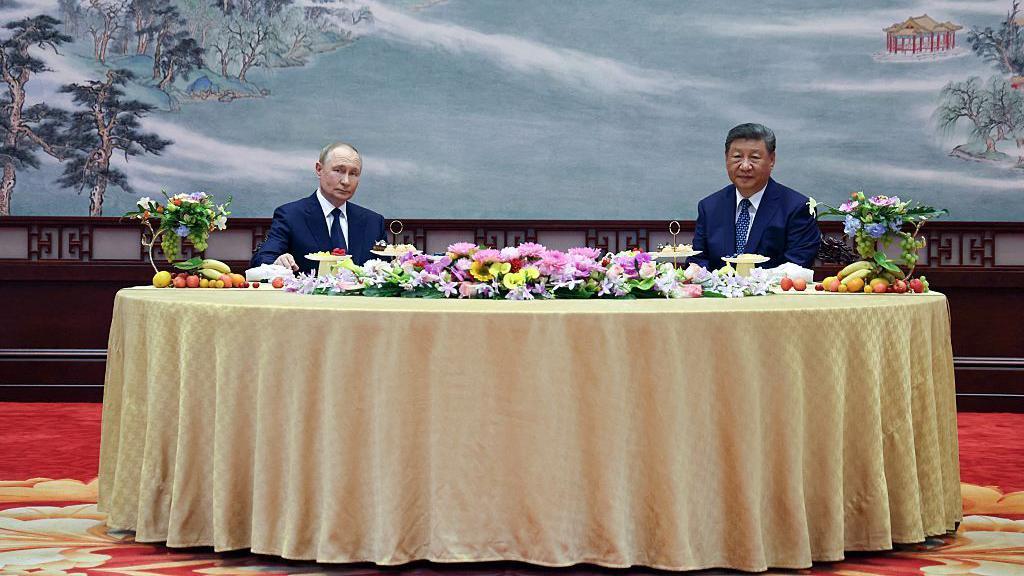 Los presidentes Putin y Xi miran a la cámara sentados en una mesa redonda con un mantel dorado y flores encima; en el fondo se ve una pintura china.