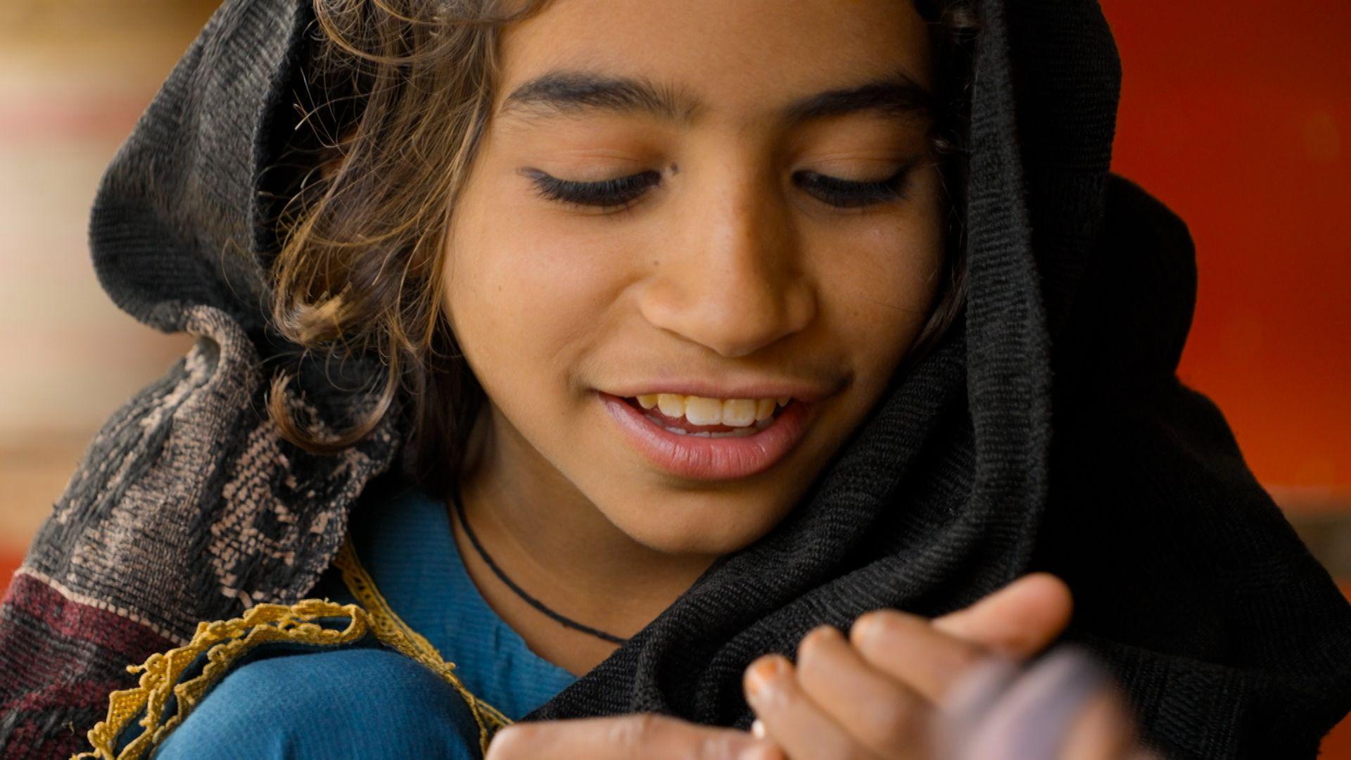 Una niña con un vestido azul sonríe, con la mirada fija en sus manos. Lleva un pañuelo oscuro con un borde estampado sobre la cabeza. 