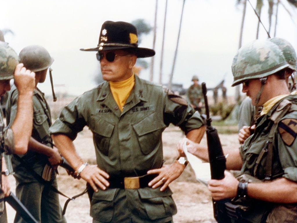 Robert Duvall interpretando al teniente coronel Kilgore, rodeado de jóvenes soldados. Duvall está parado en la mitad, con un gran sombrero negro, gafas oscuras y una pañoleta amarilla
