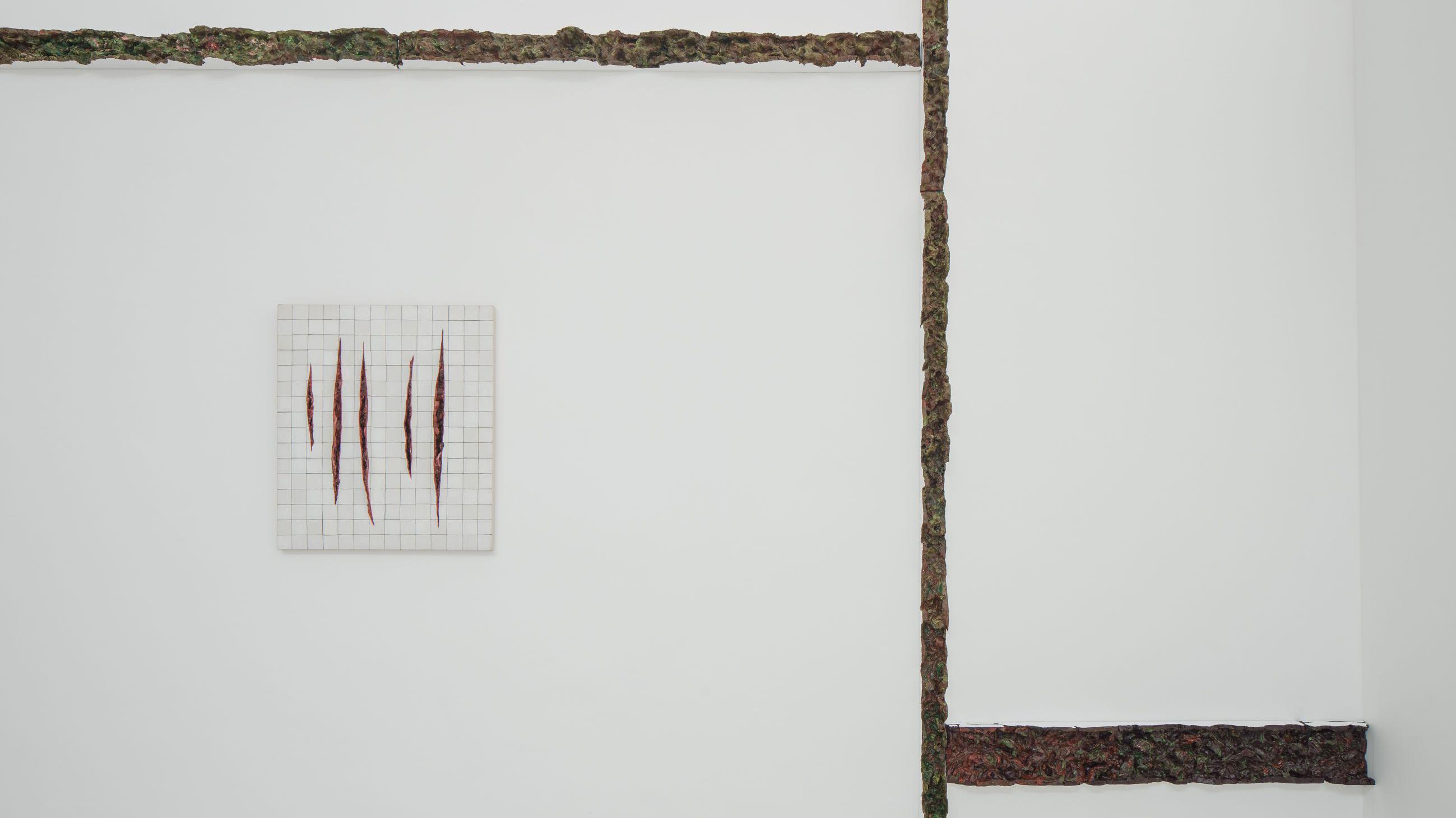 Em uma parede branca minimalista, uma pequena obra de azulejos exibe cortes verticais que lembram feridas abertas, enquanto estruturas org&acirc;nicas e escuras percorrem o espa&ccedil;o como fissuras ou cicatrizes.