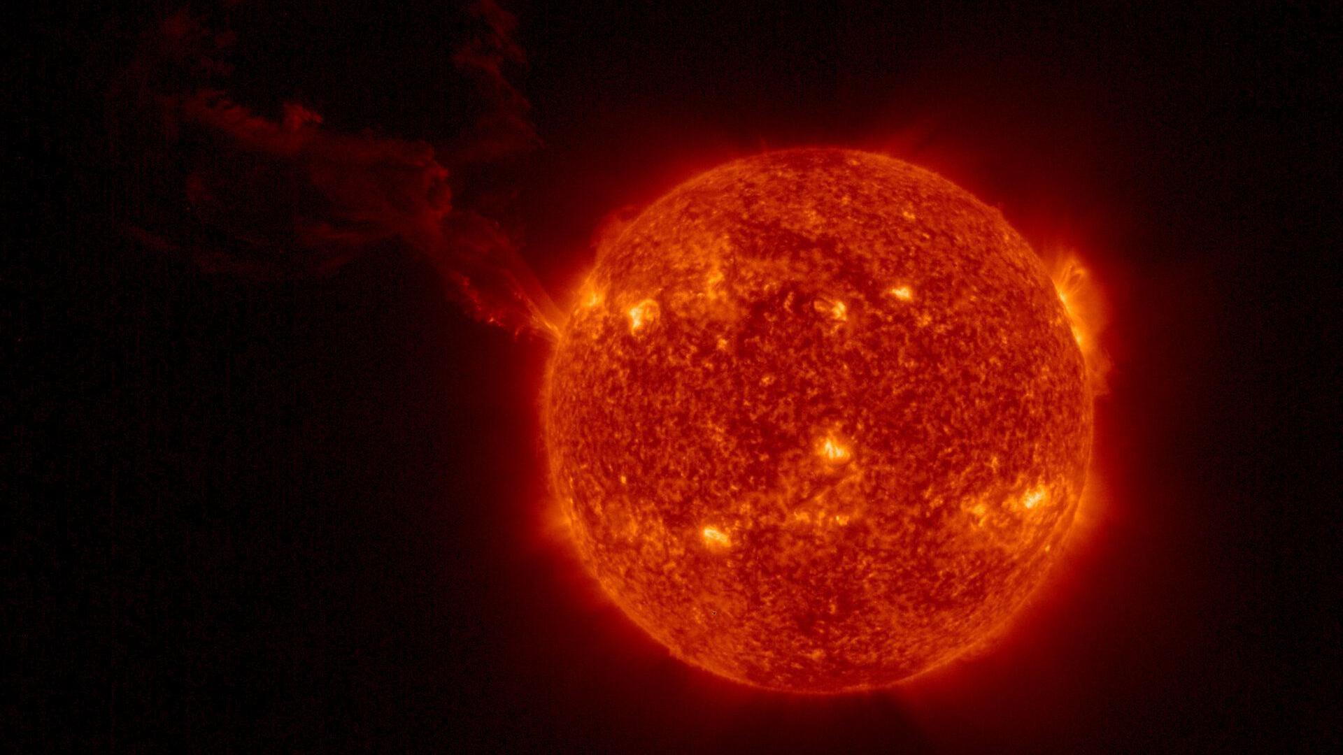 El Sol se ve como una bola roja con puntos brillantes de color amarillo en su superficie; una larga nube se extiende desde la izquierda.