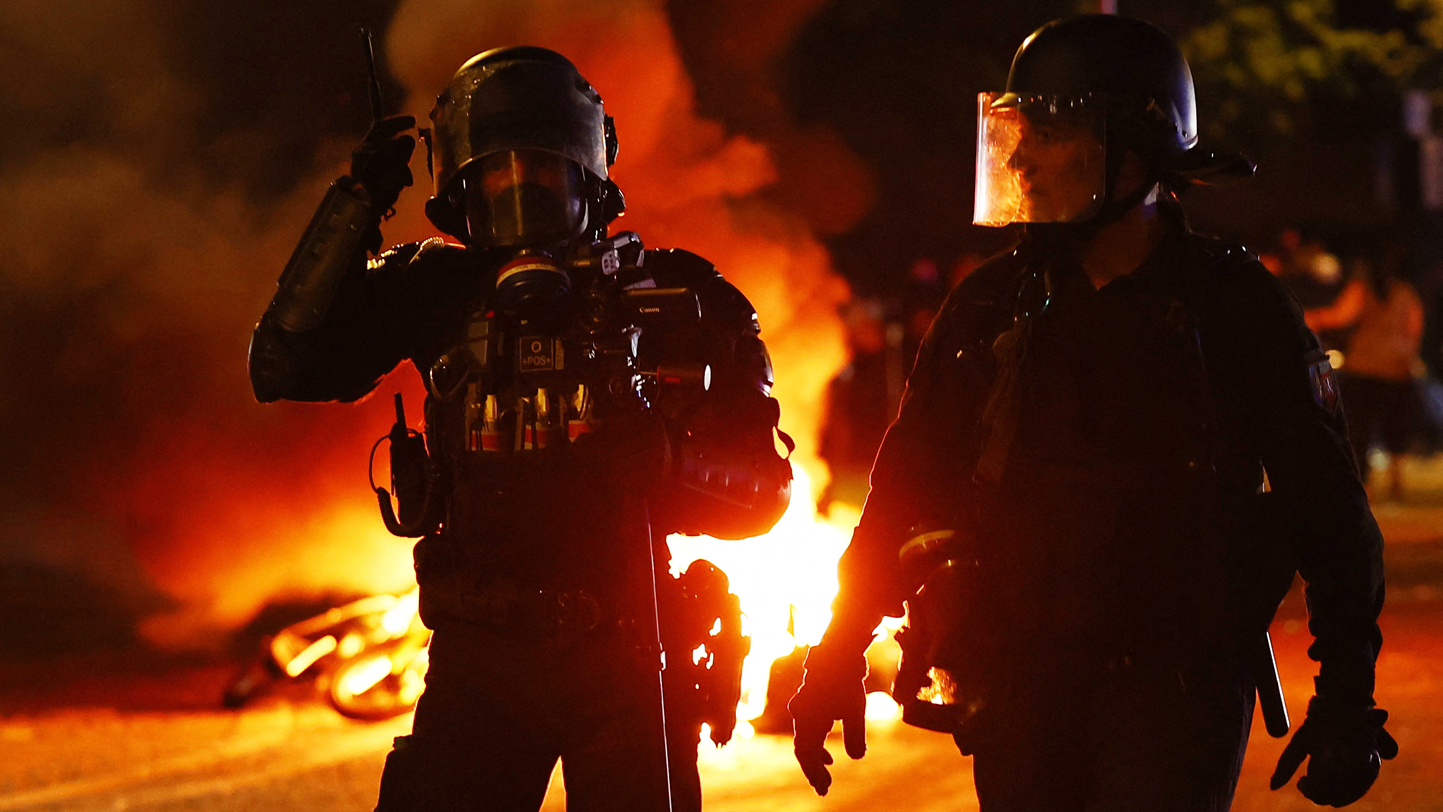 Dos policías, con guantes, cascos y mascarillas de plástico protectoras, caminan por una calle y en el fondo se fuego. 