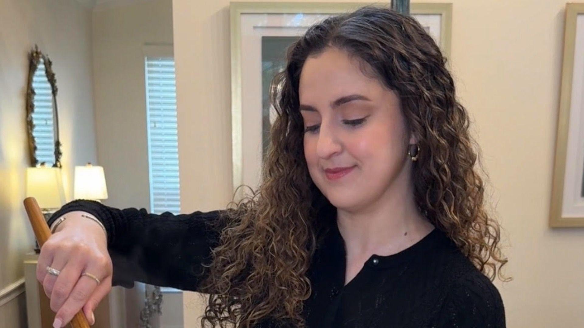 Žena u crnoj majici stoji i gleda preko kuhinjskog pulta. Ima dugu kovrdžavu kosu koja joj pada preko ramena. Drži drvenu kašiku i izgleda kao da meša hranu.