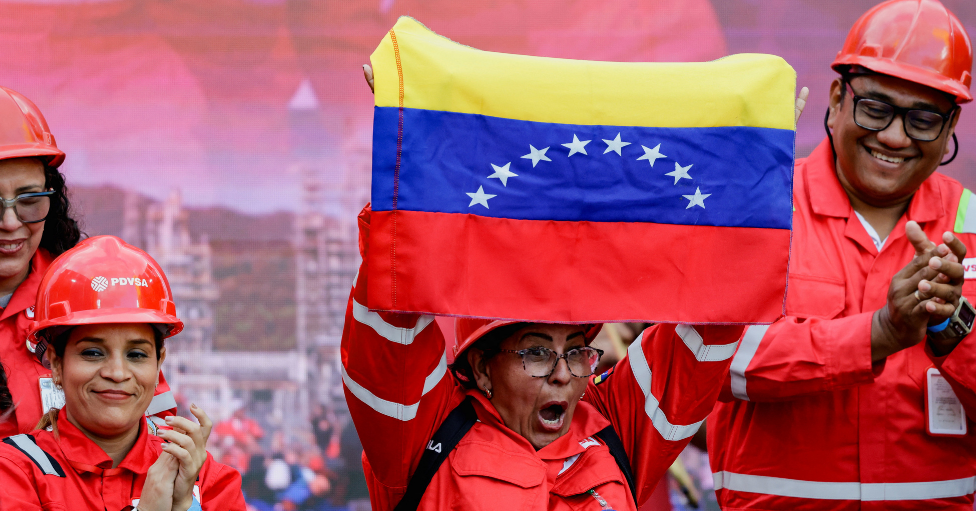 Una mujer uniformada como operaria muestra una bandera de Venezuela mientras otros trabajadores aplauden y sonríen. 