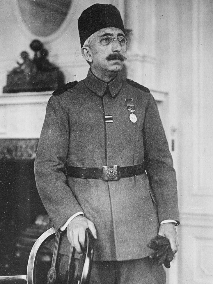Retrato del sultán Mehmed VI. Tiene puestos lentes y un tarboosh en la cabeza. Viste un uniforme, con un cinturón negro y le cuelga una medalla en el lado izquierdo de su pecho. Con una mano sostiene unos guantes negros y con la otra tiene agarrada una silla.