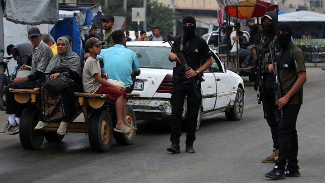 Tres milicianos con el rostro cubierto y armados con fusiles patrullan una calle en Gaza. Junto a ellos, pasa un auto blacno que tira de un carro con una mujer y un niño.