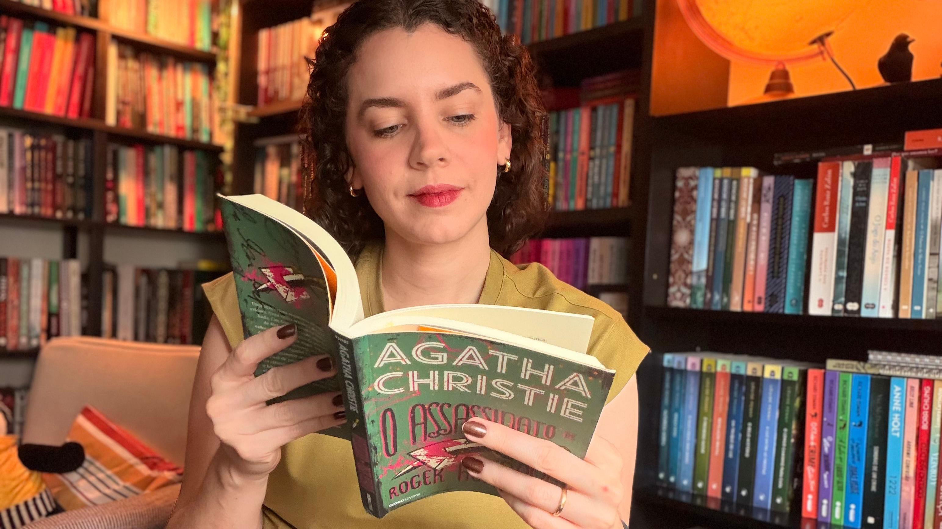 Duda Menezes, uma mulher de cabelos curtos e cacheados, l&ecirc; um livro de Agatha Christie em uma sala com estantes cheias de livros ao fundo. Ela usa blusa clara, brincos pequenos e segura o livro aberto com as duas m&atilde;os, sentada em um ambiente iluminado com luz amarelada.