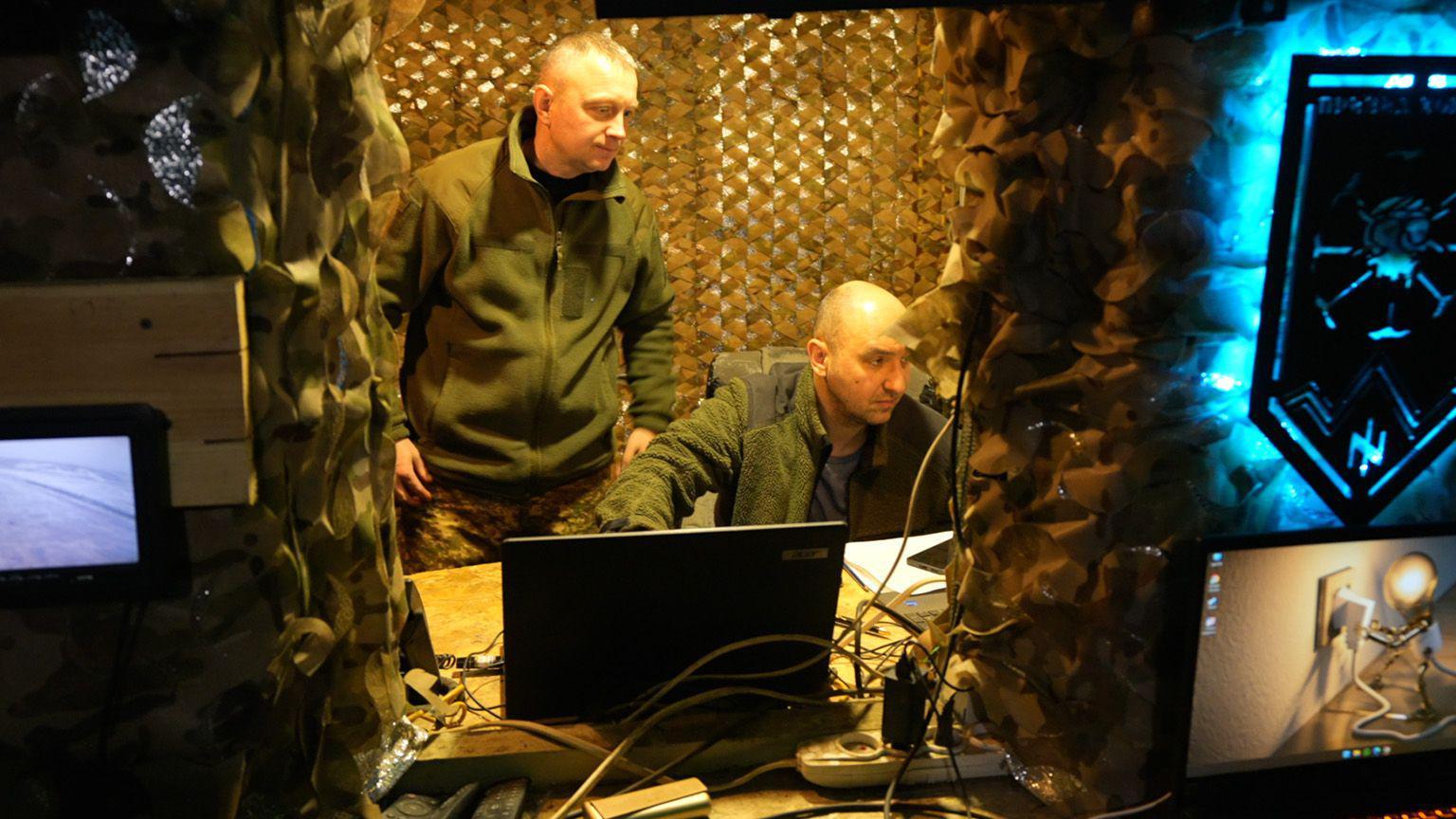 Dois homens camuflados observam equipamentos tecnol&oacute;gicos