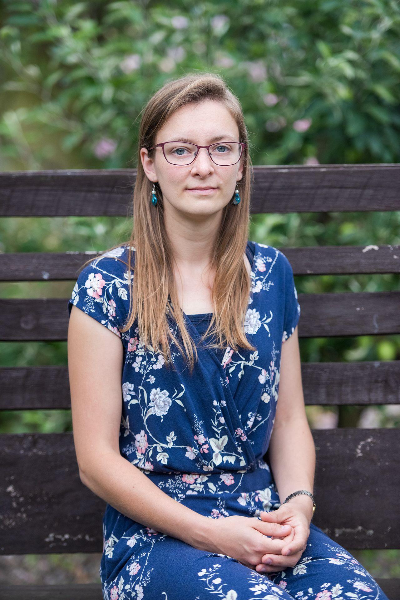 Mulher com vestido azul floral, sentada em um banco marrom, com um arbusto florido verde e rosa ao fundo