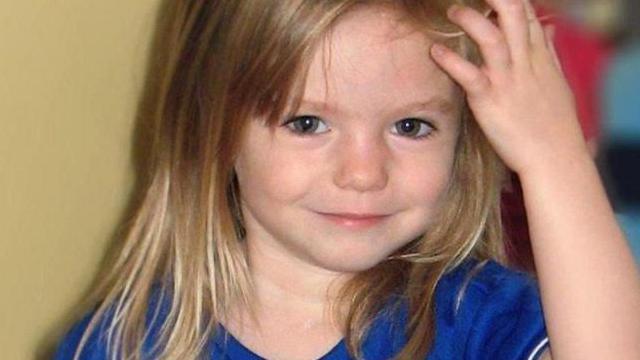 Madeleine McCann, com três anos de idade, olha para a câmera vestindo uma camisa de futebol azul e branca, com a mão esquerda levantada sobre os cabelos