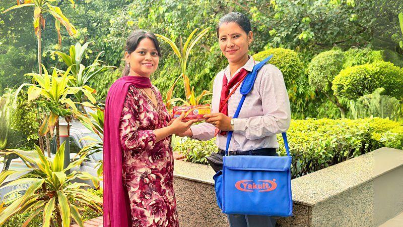 Mo&ccedil;a do Yakult faz entrega do produto para uma cliente vestida com roupas indianas