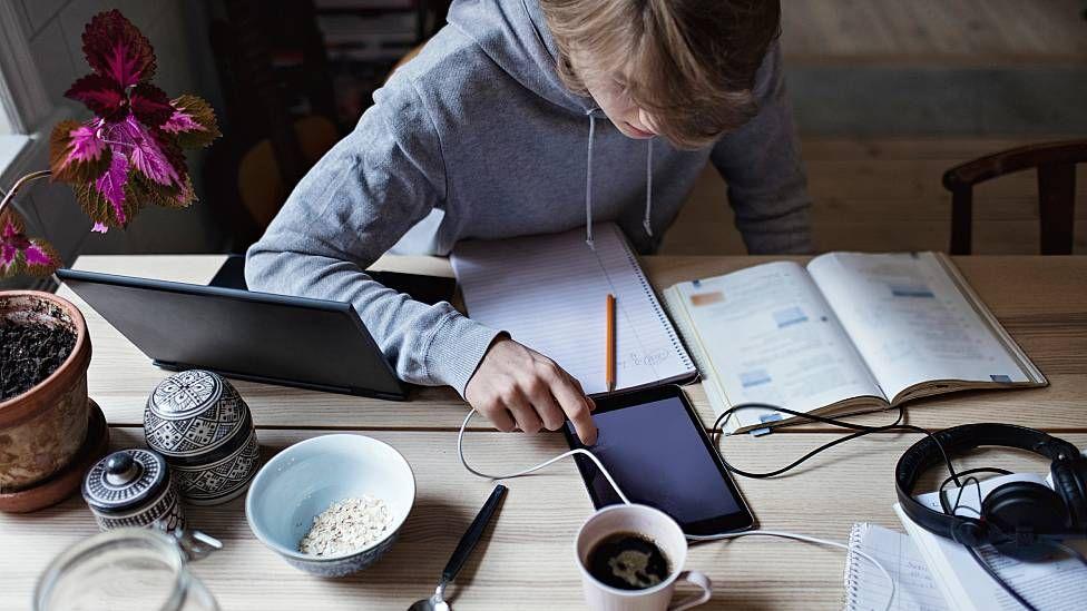Un joven estudiando sentado. En la mesa tiene un laptop, una tablet, libros y cuadernos de apuntes abiertos y una taza de café