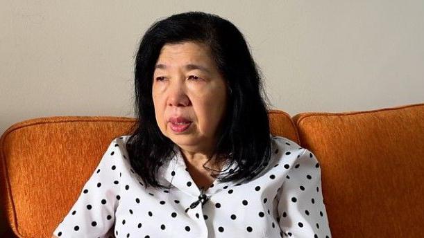 Susanna Liew, vestida con un top blanco con lunares negros, está sentada en un sofá marrón.