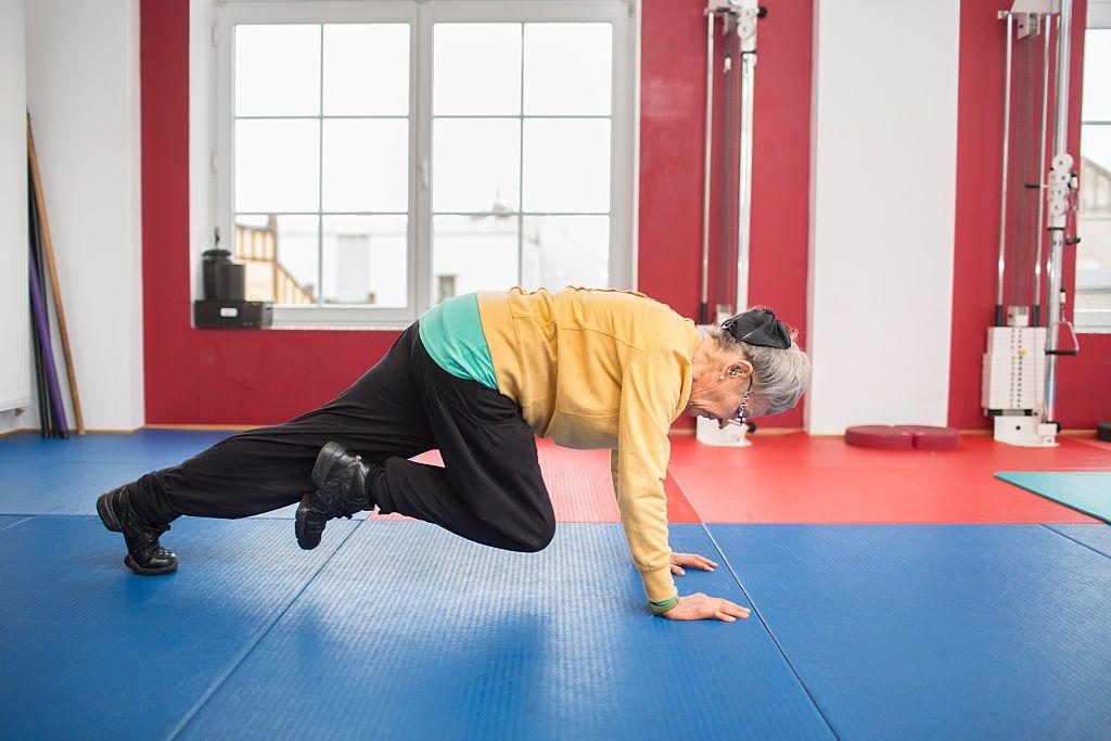 أمرأة مُسنة، ترتدي سروالاً رياضياً وحذاءً أسود رياضياً ونظارة نظر وسترة صفراء رياضية، وتمارس التمارين الرياضية في صالة ألعاب رياضية.