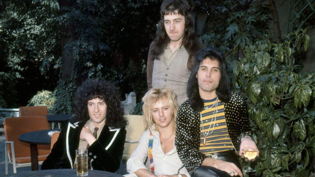Los integrantes de la banda Queen están sentados en una mesa mirando a la cámara.