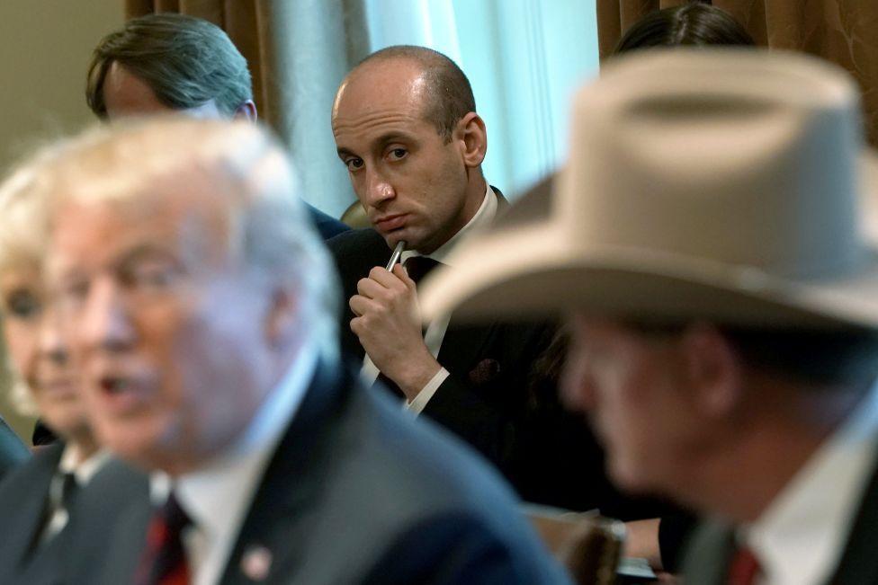 Stephen Miller aparece ao fundo, em foco n&iacute;tido. Ele segura uma caneta e observa Trump, que fala em primeiro plano e est&aacute; desfocado na imagem