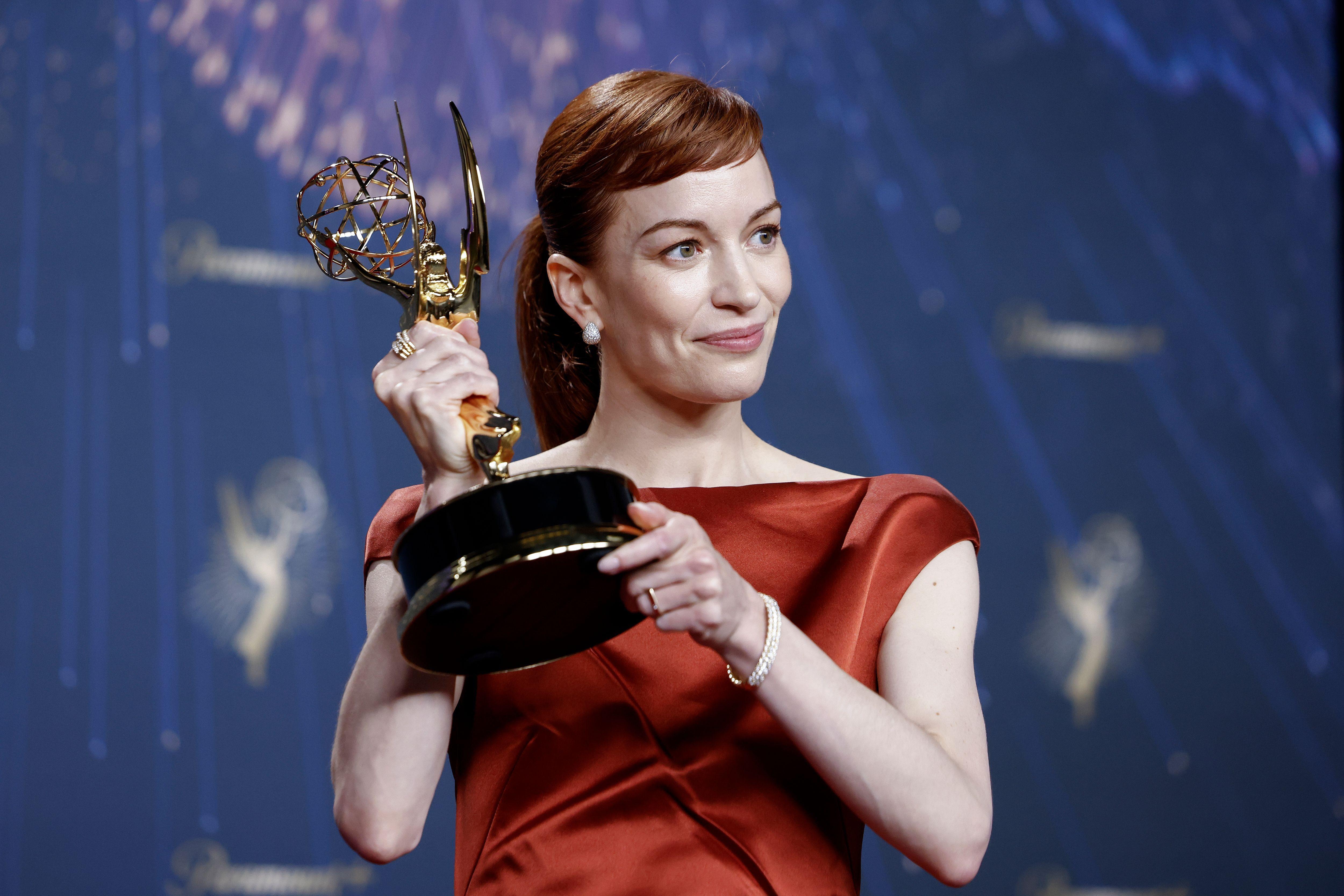 Britt Lower, con un vestido rojo de mangas cortas, sonríe mirando hacia la derecha mientras levanta la estatuilla del Emmy con ambas manos.