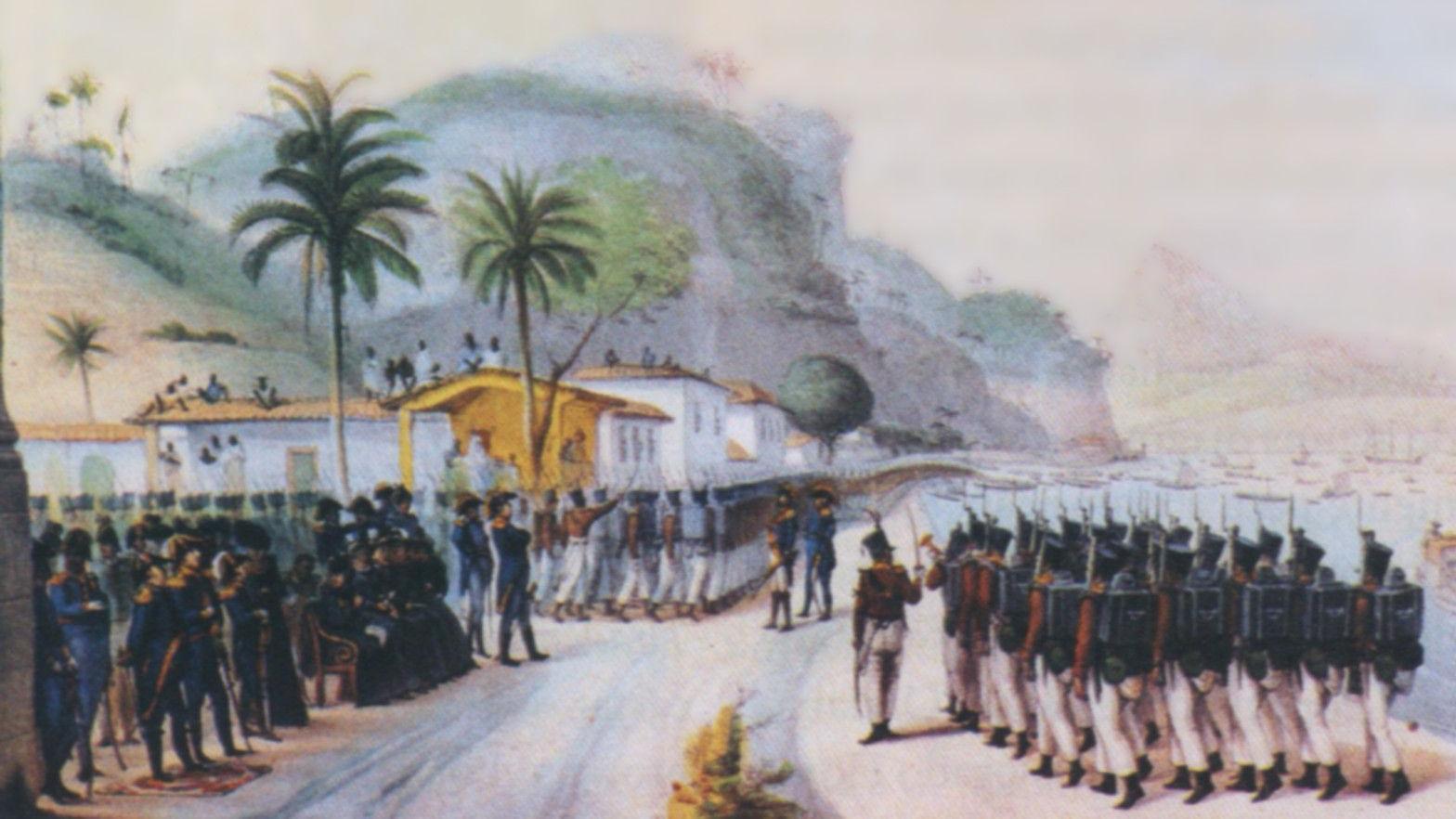 A imagem mostra uma cerim&ocirc;nia militar &agrave; beira-mar, com uma tropa de soldados em forma&ccedil;&atilde;o diante de autoridades e civis reunidos em frente a casas coloniais. Ao fundo, destacam-se morros cobertos por vegeta&ccedil;&atilde;o, palmeiras e embarca&ccedil;&otilde;es ancoradas na ba&iacute;a, compondo uma cena de car&aacute;ter hist&oacute;rico.