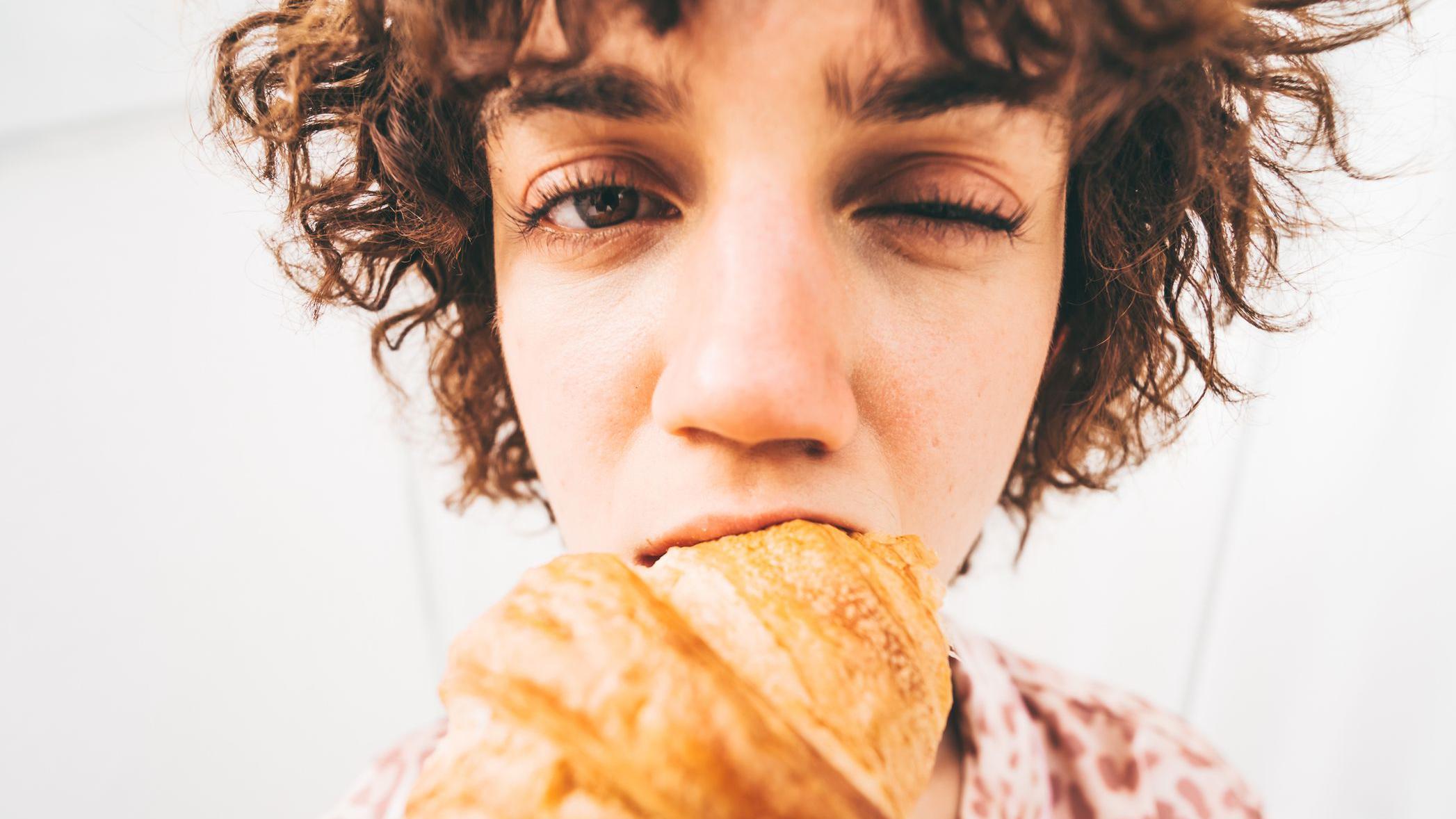 Una mujer muerde un croissant, mientras cierra un ojo. El lente ojo de pez distorsiona su cara y el croissant   