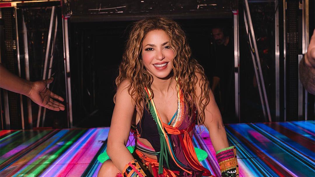 Shakira com uma roupa colorida sentada no palco. Ela sorri para a foto