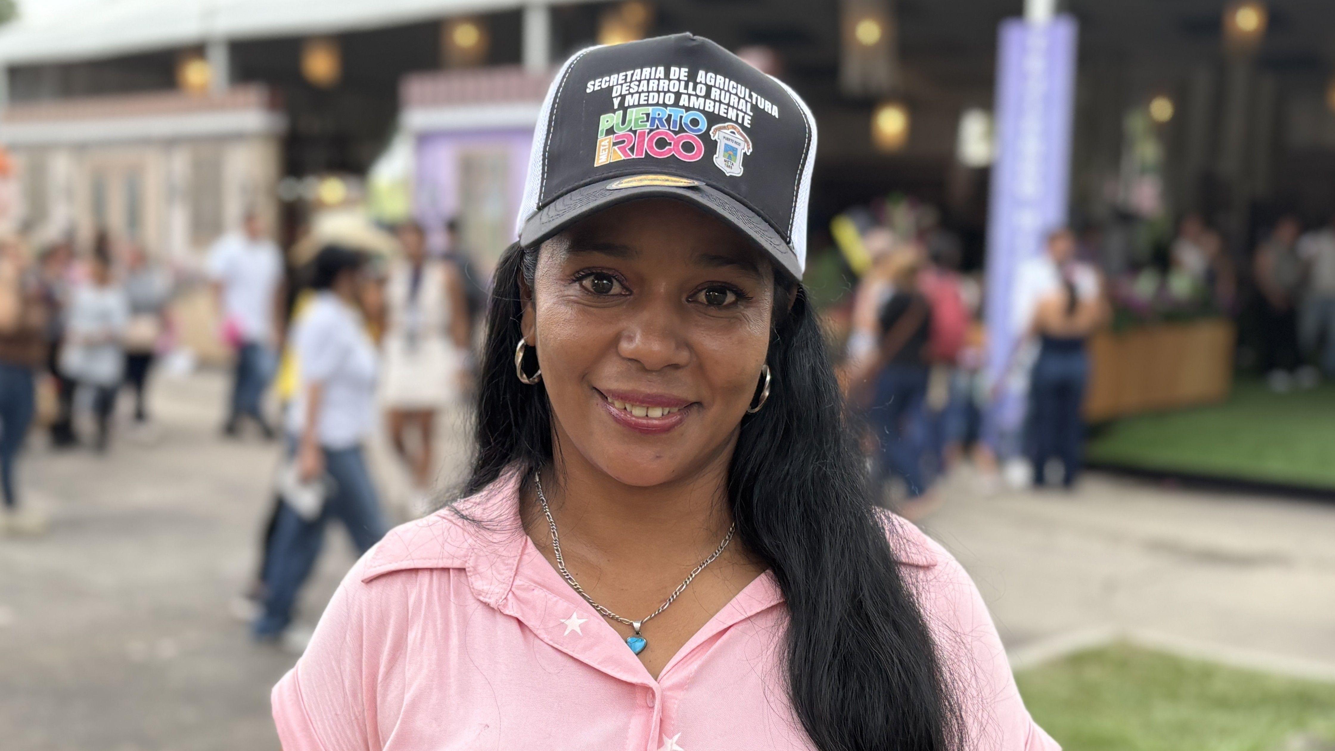 Doralba Bejarano sorri para a c&acirc;mera em uma pra&ccedil;a p&uacute;blica movimentada. Ela usa um bon&eacute; preto com frases que incluem o nome 