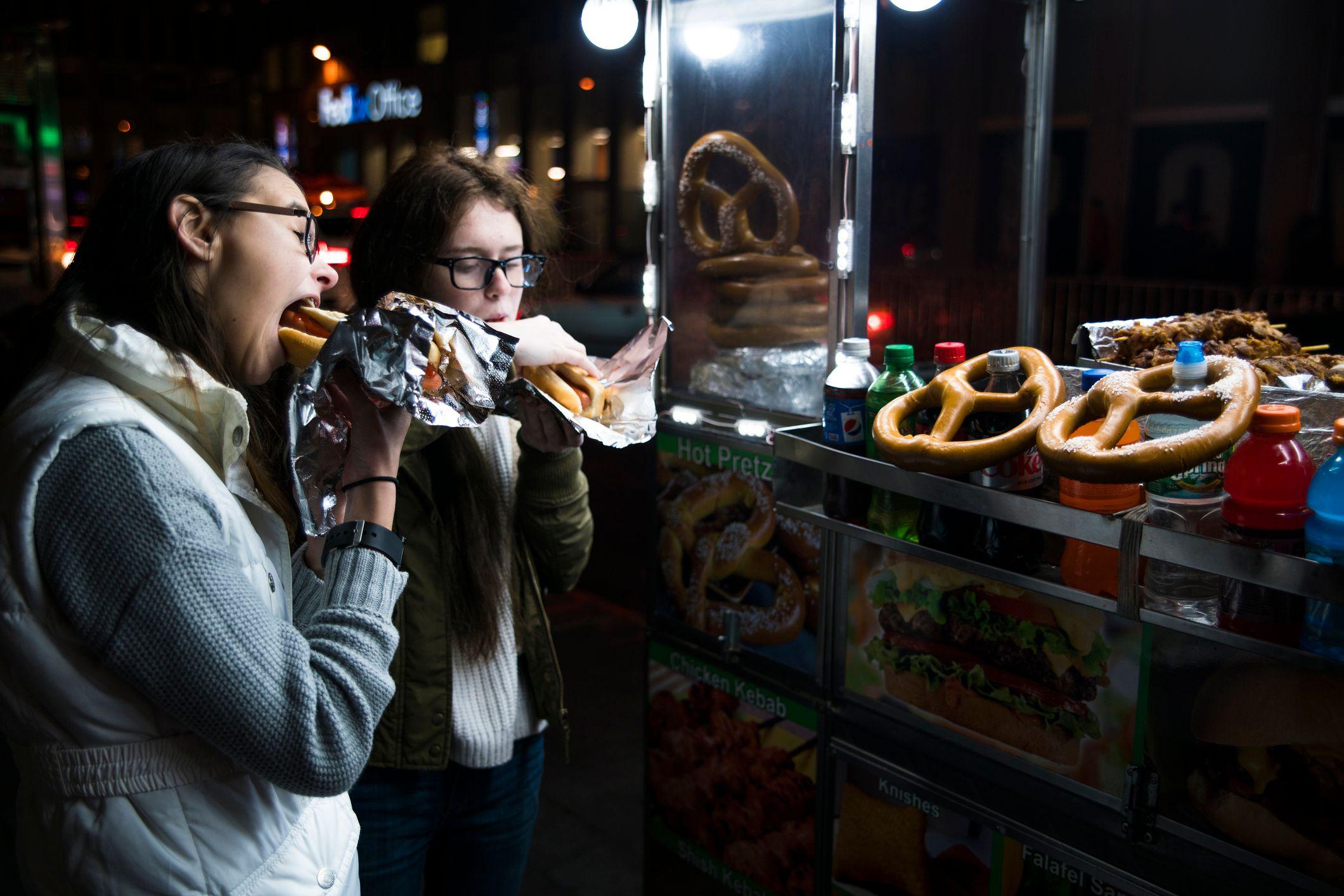 Two girls eat hot dogs outside a fast food stall. It is dark and they are both wearing jackets and jumpers.