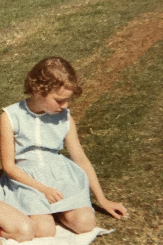 Uma fotografia colorida antiga de uma menina loira sentada em um campo gramado. A crian&ccedil;a veste um vestido azul-claro sem mangas e est&aacute; sentada sobre uma manta branca estendida na grama