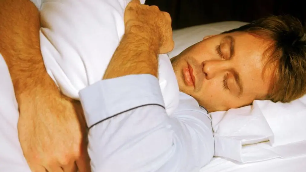 رجل ينام على سرير بمفارش بيضاء اللون ممسكاً بغطاء.