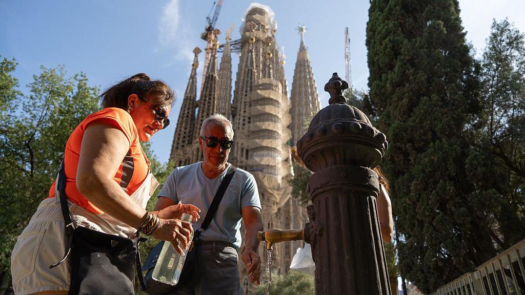 Un hombre y una mujer rellenan de agua sus botellas en una fuente pública frente a la basílica de la Sagrada Familia en Barcelona el 27 de junio de 2025, un día antes de que se espere oficialmente que comience la primera ola de calor del año.